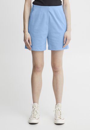 Pantaloni corti azzurri su misura con pieghe, tasche laterali e una texture liscia, abbinati a sneakers bianche alte.