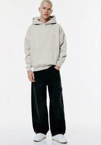 Felpa grigia in cotone con tasca a marsupio, abbinata a jeans neri larghi. Sneakers bianche completano l'outfit. Il modello è in piedi su uno sfondo neutro.
