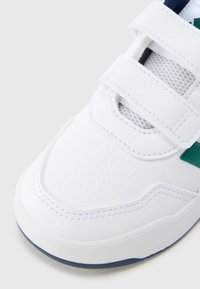 Biały sneaker z teksturowanym syntetycznym cholewką, paski na rzepy, zielony akcent i zaokrąglony nosek. Charakteryzuje się kontrastową niebieską gumową podeszwą.