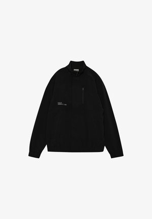 Chaqueta cortavientos negra con un cuello alto, cremallera frontal y bolsillos, que presenta un pequeño texto detallado en el lado izquierdo.