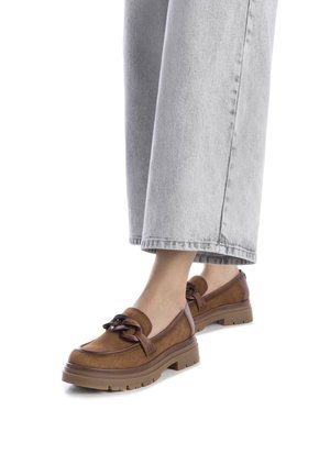 Mocasines de ante marrón con suela de goma gruesa, que cuentan con una correa decorativa de cuero marrón y un acabado suave, combinados con unos pantalones de mezclilla de pierna ancha en gris claro.