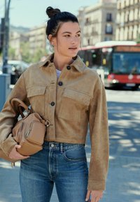 Giacca in suede marrone cropped con chiusura a bottone e due tasche frontali, abbinata a jeans a vita alta azzurri e una borsa beige.