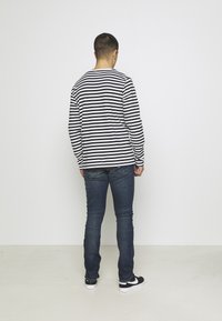 Černobíle pruhované tričko s dlouhým rukávem, modré džíny, černo-bílé tenisky. Osoba otočená zády, ukazuje ležérní vrstvený styl oblečení.