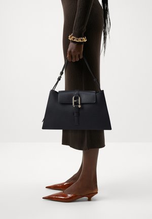 Sac à main en cuir noir au design structuré, doté d'une anse, de quincaillerie dorée et d'un détail de fermoir. Le modèle porte une robe marron côtelée et des talons marron.