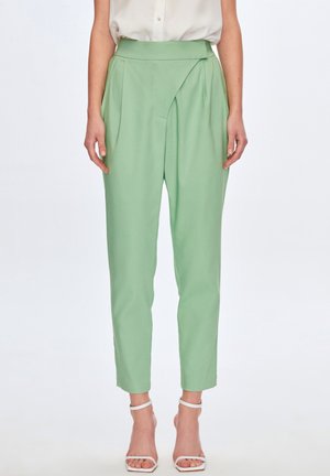 Kvinde iført højtaljede, lysegrønne, tilspidsede bukser med læg, kombineret med hvide remsandaler og en indstoppet bluse.