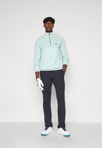 Camisola com fecho éclair verde menta com logo preto, calças escuras, sapatos brancos com detalhes azuis e um boné preto. Modelo a usar luva de golfe.