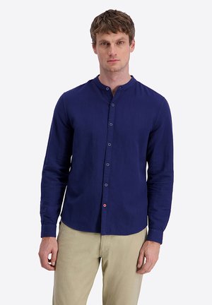 Man wearing a navy blue mandarin collar button-up shirt and beige pants, standing against a plain light background.