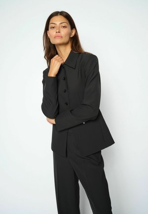 Donna con capelli lunghi e scuri che indossa una giacca nera con bottoni e pantaloni abbinati, posando con una mano vicino al collo su uno sfondo uniforme.