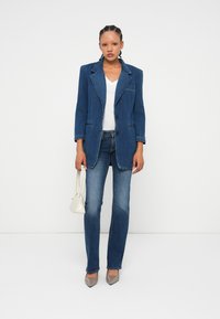 Denim blazer v temno modri barvi z oblikovanimi rameni, v kombinaciji z belo s V-izrezom, svetlo modrih kavbojskih hlačah in sivih čevljih s koničastim nosom.