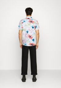 Lyhythihainen kukallinen paita pastelliväreissä, joissa on sinisiä, vaaleanpunaisia ja vihreitä kuvioita; yhdistettynä mustiin housuihin ja mustiin jalkineisiin.