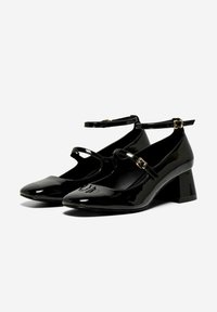 Chaussures Mary Jane en verni noir avec un bout pointu, une sangle au niveau de la cheville et un talon épais. Texture lisse, avec de petites découpes et des éléments en métal.