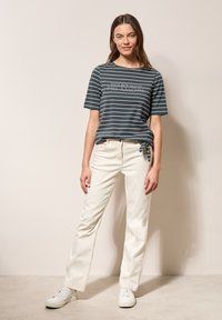 Camiseta de manga corta a rayas en gris oscuro y blanco, con detalle de texto; combinada con jeans de pierna recta en blanco roto y zapatillas blancas.