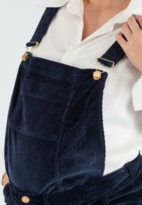 Navy corduroy salopette met verstelbare schouderbanden, gouden hardware, vo zak en textuurstof, gedragen over een wit shirt.