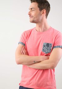 Camiseta de manga corta rosa hecha de tela suave, con un bolsillo con estampado y puños de manga con un patrón contrastante. Vista frontal.