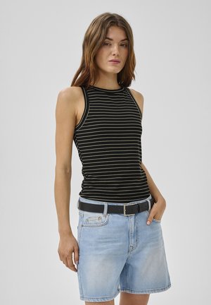 Kobieta ubrana w czarno-biały, bezrękawnik w paski oraz jasnoniebieskie szorty jeansowe z czarnym paskiem, stojąca z jedną ręką w kieszeni.