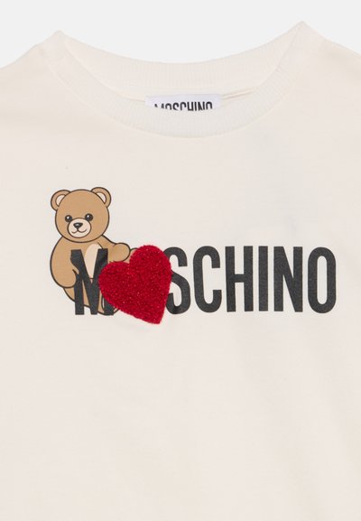 Бяла риза с черен текст "MOSCHINO," кафяво плюшено мече, облегнато на буквата "M,", и текстурирано червено сърце над буквата "O."