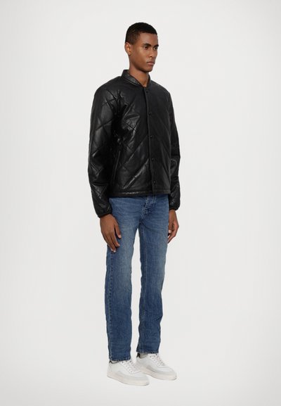 Veste en cuir noir matelassée avec une fermeture à bouton pression, portée sur un jean bleu et des baskets blanches. Dotée d'un col arrondi et d'une surface texturée.