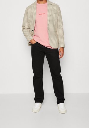 Osoba v béžovém kostkovaném saku, růžové košili s textem "DOLCE VITA", černých kalhotách a bílých teniskách stojí s jednou rukou v kapse.