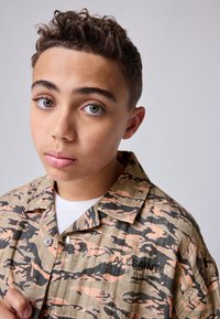 Overhemd met een camouflagepatroon in groen, zwart en oranje, korte mouwen en een knoopsluiting. Heeft een "All Saints" logo.