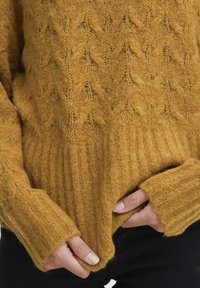 Gestrickter senfgelber Pullover mit einem strukturierten Rautenmuster und gerippten Bündchen. Weiches Material mit einem leicht lockeren Sitz, die Finger greifen den Saum.