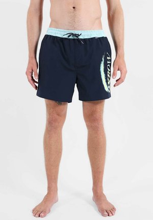 Homme portant un short de bain bleu marine avec une ceinture bleu clair et un logo blanc « Kappa » sur la jambe droite, debout pieds nus sur fond blanc.