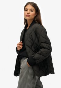 Zwarte gewatteerde bomberjack met ribbelboorden en kraag. Voorzien van een ritsluiting en logo detail op de mouw, gestyled over gestreepte broek.