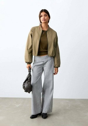 Olivgrüne Bomberjacke, dunkelgrüner Pullover, weit geschnittene hellgraue Jeans, schwarze Stiefeletten und schwarze Handtasche in einem minimalistischen Ambiente.