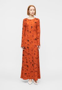 Γυναίκα που φοράει ένα πορτοκαλί φόρεμα με μακρύ μανίκι, μήκος μέχρι το πάτωμα, με σκοτεινά λουλουδένια σχέδια, στέκεται μπροστά σε ένα απλό ανοιχτό φόντο.