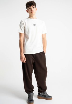 Młody mężczyzna stoi z rękami w kieszeniach, ubrany w białą koszulkę z krótkim rękawem z małym emblematem na piersi, czarne szerokie spodnie i szare tenisówki.