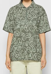 Polo a maniche corte in tessuto verde oliva, con scollo con zip e motivo astratto ripetuto in verde chiaro. Include una tasca sul petto.