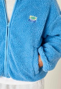 Blauwe fleecejas met een ritsluiting, textiele stof en een fleece-achtige uitstraling. Voorzien van een klein merklogo op de borst.