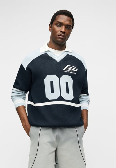 Άνδρας που φοράει μπλε και γαλάζιο πουλόβερ με τον αριθμό 00 και το λογότυπο "Off-White", σε συνδυασμό με ανοιχτόγκρι παντελόνια σε λευκό φόντο.