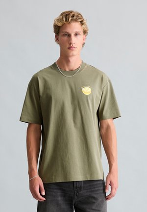 Jonge man met blond haar draagt een olijfgroen T-shirt met een kleine gele citroengrafiek en zwarte jeans tegen een lichtgrijze achtergrond.