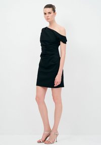 Schwarzes schulterfreies Kleid mit geraffter Textur, figurbetontem Schnitt und mini Länge. Das Model trägt silberne, strappy High Heels. Glatte Stoffoberfläche.