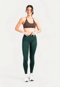 Brassière de sport marron et leggings taille haute vert foncé, tous deux en tissu extensible. Le modèle porte des baskets blanches et se tient les mains sur les hanches.