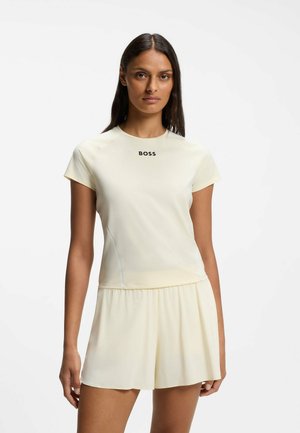 Kvinde med langt mørkt hår har en cremefarvet kortærmet top med teksten "BOSS" og matchende løse shorts mod en ensfarvet hvid baggrund.