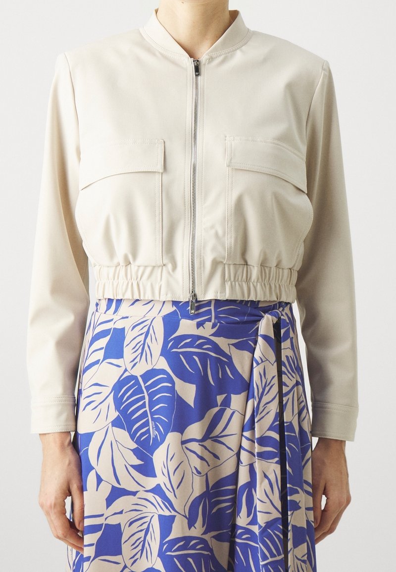 Femme portant une veste courte crème zippée avec des poches à rabat et une jupe à motif feuilles bleu et beige nouée à la taille.