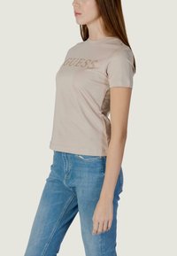 Camiseta de manga corta beige con un logo brillante "GUESS" en el pecho. La tela es suave, con un diseño ajustado y cuello redondo.