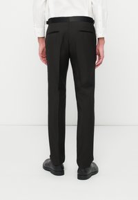 Homme portant un pantalon noir sur mesure et des chaussures de ville noires, vu de dos contre un fond blanc uni.