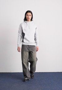 Hallikas kapuutsiga särk eesmise tasku ja nööriga, koos vaba lõikega tuhmide hallide teksade ja mustade tossudega, seismas sinisel vaibal.