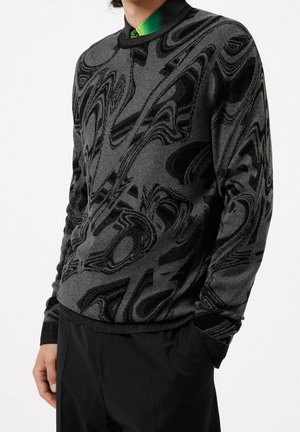 Mężczyzna ubrany w czarno-szary sweter z abstrakcyjnym wzorem, na zielonej koszuli z kołnierzykiem, czarne spodnie, ręce częściowo w kieszeniach.