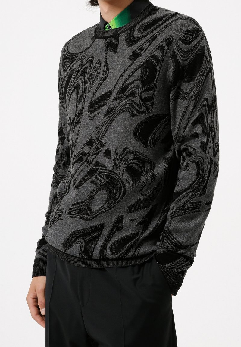 Homme portant un pull à motif abstrait noir et gris sur une chemise verte à col, et un pantalon noir, les mains partiellement dans les poches.