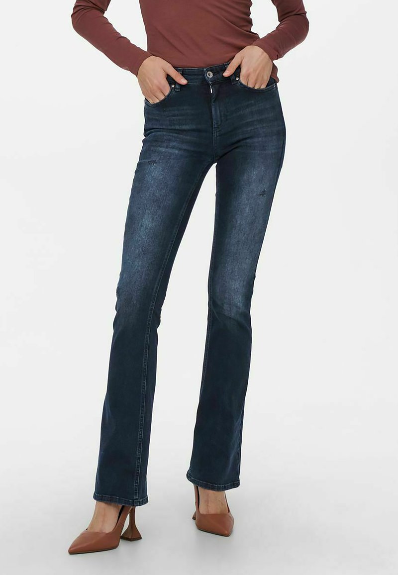 Jeans svasati in denim blu scuro con vita media, leggera scoloritura e design a cinque tasche, abbinati a scarpe con tacco marroni.