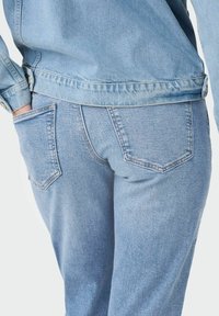 Svijetloplava traper jakna u kombinaciji sa svijetloplavim trapericama. Jakna ima zatvaranje na dugmad, dok traperice imaju dva stražnja džepa.