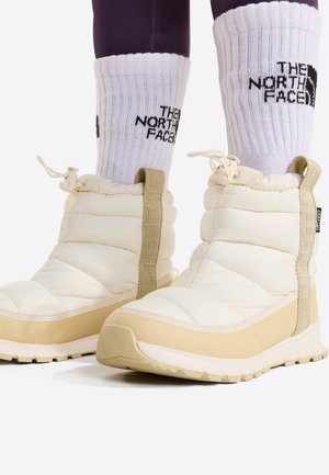 Bottes d'hiver isolantes couleur crème portées avec des chaussettes blanches The North Face et des leggings foncés sur fond blanc.