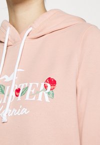 Bluza z kapturem w kolorze brzoskwiniowym z białymi sznurkami, kwiatowym nadrukiem w odcieniach czerwieni i zieleni oraz dużymi literami na przodzie. Miękki bawełniany materiał.