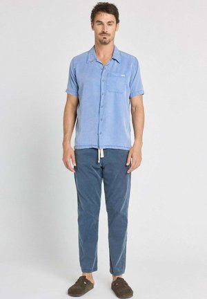 Homme debout devant un fond uni portant une chemise bleu clair à manches courtes boutonnée, un pantalon bleu foncé à cordon et des chaussons marron.