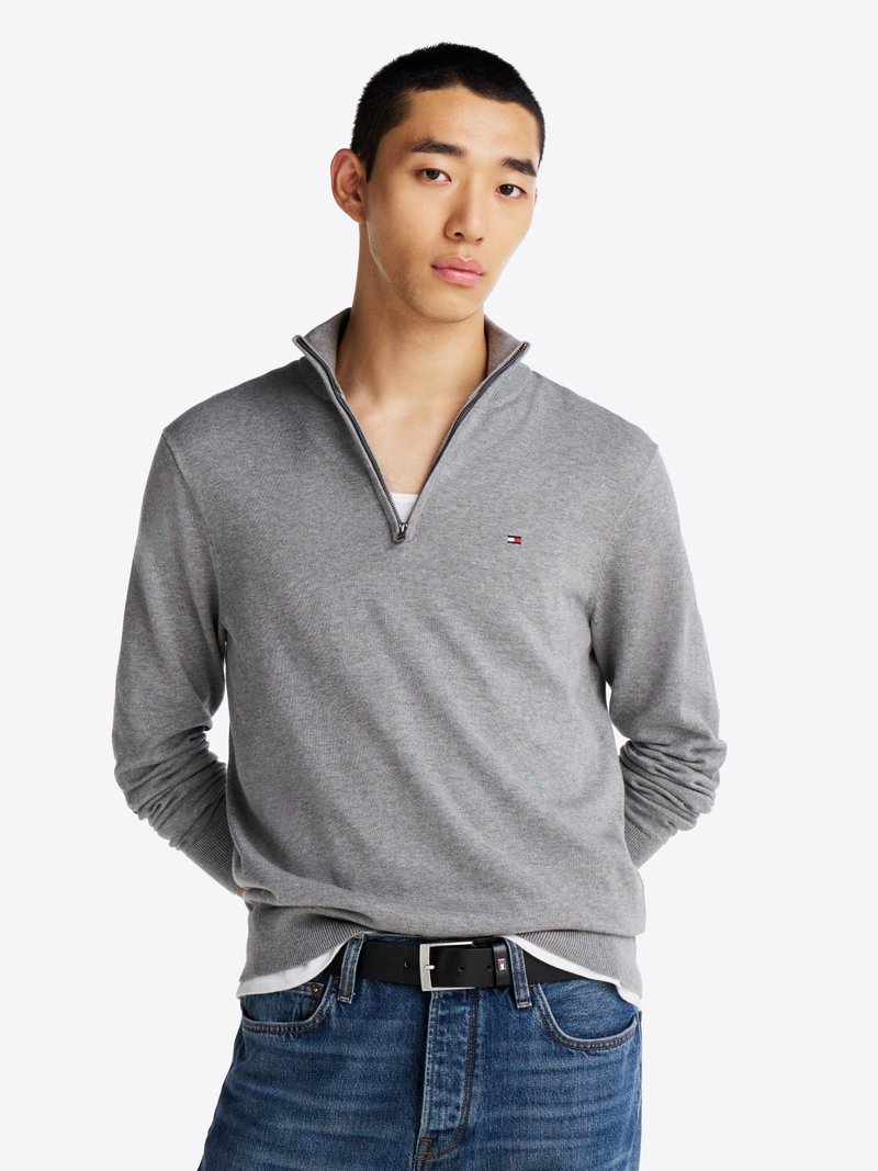GrauZip-Up-Pullover mit geripptem Kragen. Flachstrick-Textur, Material aus Baumwollmischung. Kombiniert mit blauen Jeans und einem schwarzen Gürtel.