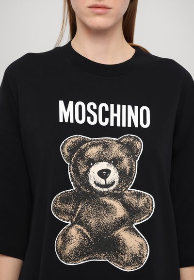 Svart t-shirt med kort ärm som har en stor grafisk bild av en nallebjörn i brunt med vita detaljer. Varumärket "MOSCHINO" är tryckt ovanför grafiken.