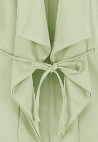 Lichtgroene wikkeltop met een gedrapeerde kraag en striksluiting, voorzien van een zachte stof en een gladde textuur, met minimale stikdetails zichtbaar.